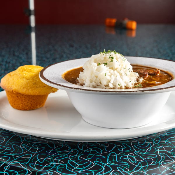 A bowl of Miss Delta's gumbo with rice and a cornbread muffin on a retro-patterned table.