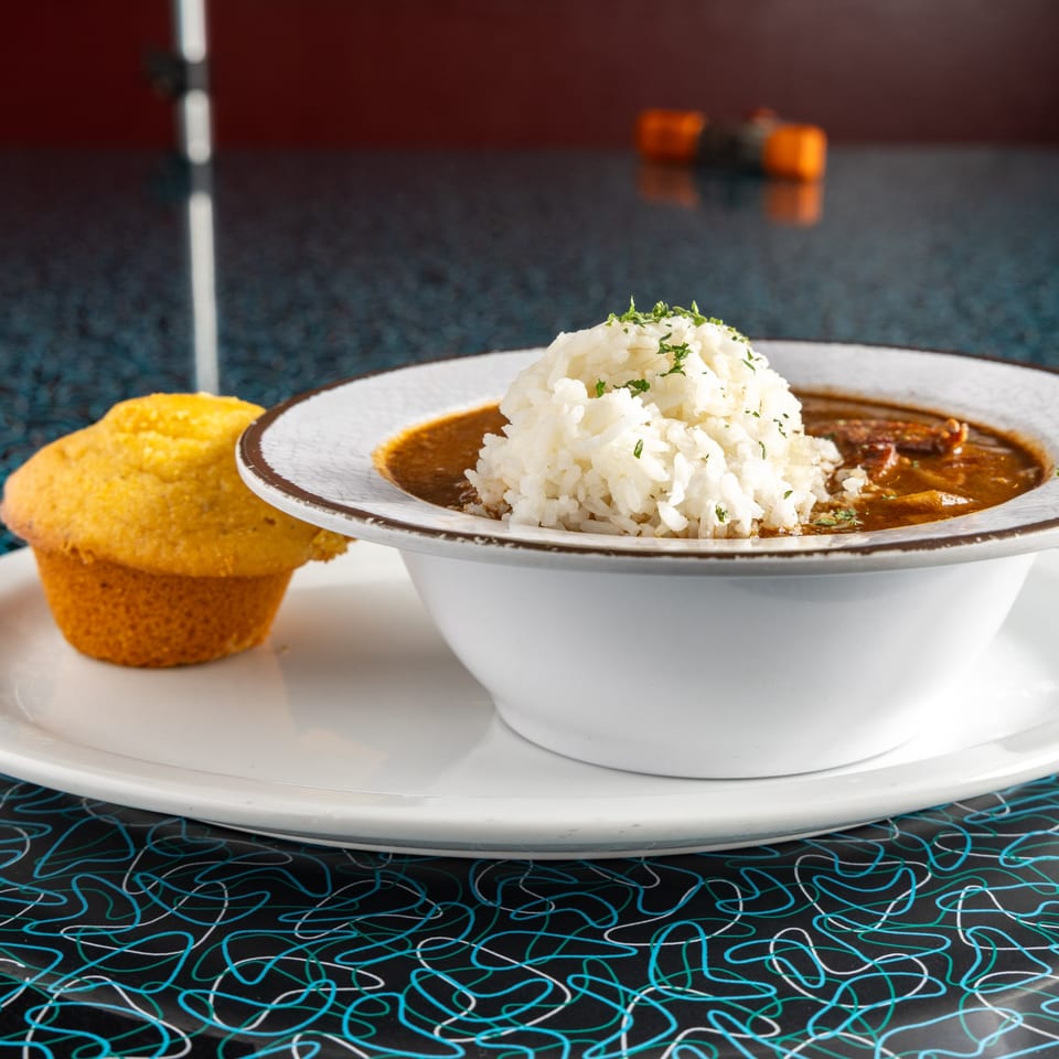 A bowl of Miss Delta's gumbo with rice and a cornbread muffin on a retro-patterned table.