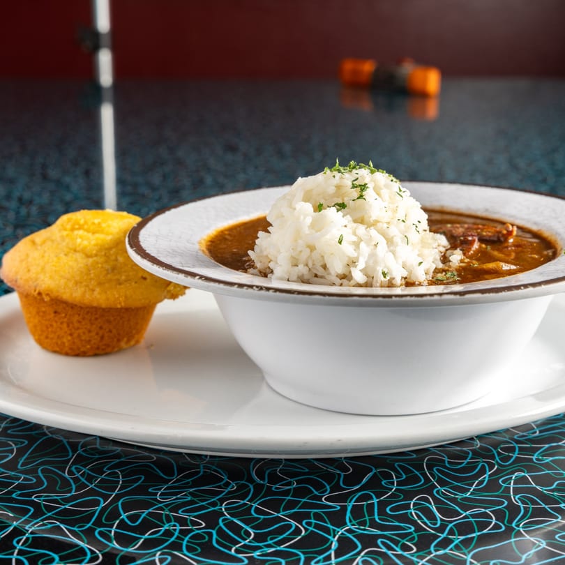 A bowl of Miss Delta's gumbo with rice and a cornbread muffin on a retro-patterned table.