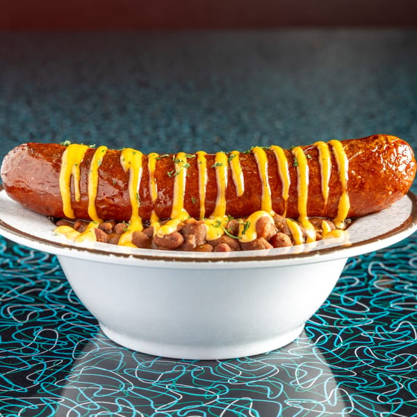 A large, grilled sausage drizzled with yellow mustard and herbs, served over beans in a white bowl.