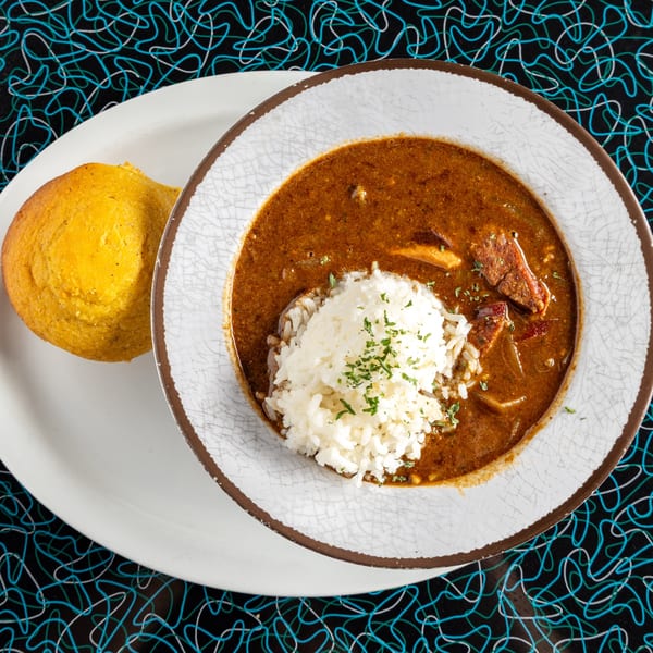 Miss Delta's gumbo with rice and cornbread, served on a white plate with a patterned background.