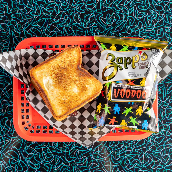 A grilled sandwich and a bag of Zapp's Voodoo chips in a red basket on a retro patterned table.