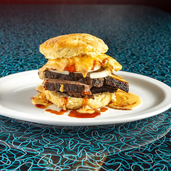 A Miss Delta BBQ brisket biscuit sandwich with melted cheese, onions, and gravy on a white plate.