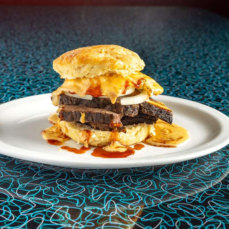 A Miss Delta BBQ brisket biscuit sandwich with melted cheese, onions, and gravy on a white plate.